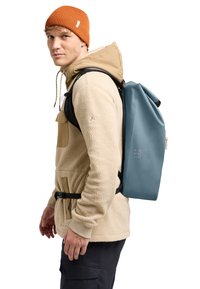 Sac à dos à rabat en matériau bleu doux et texturé. Sangles et accents noirs. Le modèle porte une veste en polaire beige et un bonnet orange.