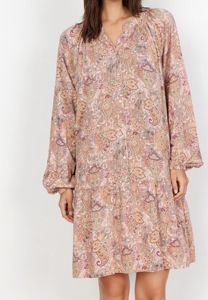 Kvinde iført en langærmet, knaplukket, knælængde kjole med pink og beige blomstrede paisley-mønstre.