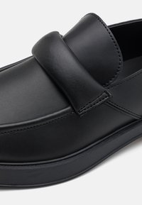 Mocassins en cuir noir avec un bout arrondi, dotés d'une lanière supérieure proéminente et de coutures visibles. Texture lisse avec une semelle plate.
