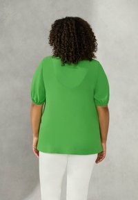 Blusa verde con maniche a sbuffo, texture liscia e scollatura arrotondata sul retro. Abbinata a pantaloni bianchi su uno sfondo neutro.
