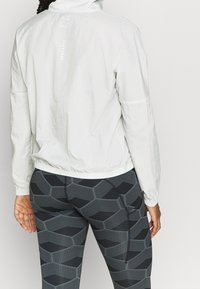 Veste blanche légère avec capuche, poignets élastiques et un motif texturé dans le dos. Associée à des leggings noirs à imprimé géométrique.