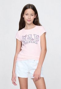 Rosa bomull t-skjorte med korte ermer, med "GAP" i marineblått med blomsterdetaljer, kombinert med hvite denimshorts. Enkel, casual design.