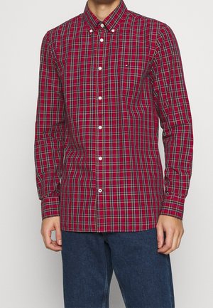 Mann trägt ein rotes und grünes kariertes Hemd mit Knöpfen und blaue Jeans, steht vor einem schlichten hellen Hintergrund.