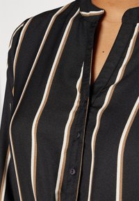 Svart blus med vertikala ränder i brunt och vitt. Tyget är mjukt, med en kraglös design och knappstängning vid nacken.