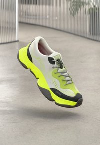 Lys grå og neon gul sneakers med svart såle og lisser, svevende over et betonggulv i en innendørs setting.