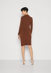 Robe ajustée à manches longues marron avec un col, présentant un design sans coutures et une longueur mi-mollet. Le modèle porte des chaussures à talons hauts blanches.