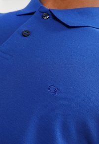 Blå polo skjorta med två svarta knappar och en liten broderad "ck"-logotyp på bröstet.