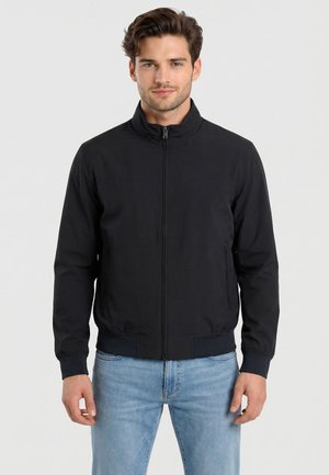 Jeune homme portant une veste noire zippée et un jean bleu clair debout devant un fond blanc uni.