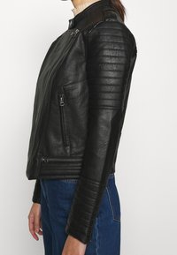 Giacca da moto in pelle nera con dettagli trapuntati sulle spalle e sulle maniche, zip frontale asimmetrica e tasche laterali. Texture liscia.