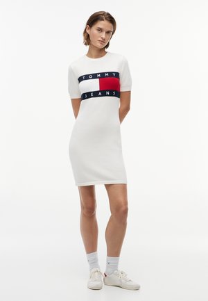 Młoda kobieta ubrana w białą sukienkę z krótkim rękawem z logo Tommy Jeans, białe skarpetki i białe trampki, stojąca na tle jednolitego białego tła.