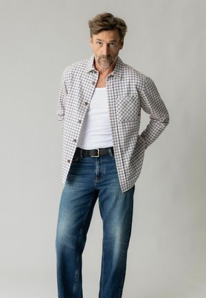 Mand med kort brunt hår og skæg står med hænderne bag ryggen iført en hvid tanktop, ternet skjorte med knapper, blå jeans og sort bælte.