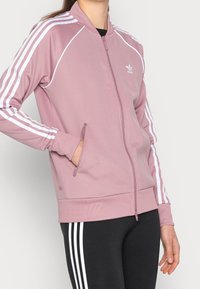 Person som bär en mauve Adidas zip-up jacka med vita ränder på ärmarna och svarta byxor med vita ränder, hand i jackfickan.