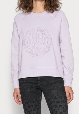 Žena nosí levandulovou mikinu s kruhovým vystouplým logem "MOSH FASHION" a tmavé vzorované džíny, stojící před neutrálním pozadím.