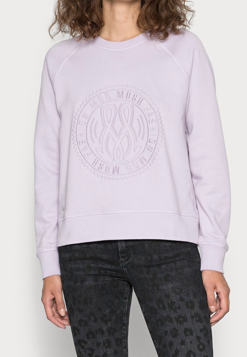 Femme portant un sweat lavande avec un logo circulaire embossé "MOSH FASHION" et un jean foncé à motifs, debout devant un fond neutre.