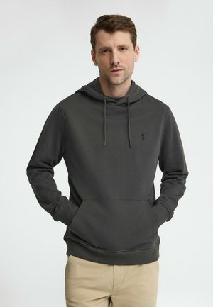 Uomo con pelle chiara e capelli castani corti, che indossa una felpa con cappuccio grigio scuro e pantaloni beige, in piedi con le mani nelle tasche contro uno sfondo neutro.