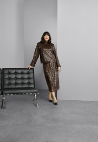 Bruine pailletten top met hoge hals en losse pasvorm, gecombineerd met een bijpassende pailletten rok. Zwarte puntige flats en een minimalistische zwarte stoel in de buurt.