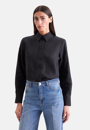 Femme portant une chemise noire boutonnée rentrée dans un jean bleu taille haute, debout les mains détendues le long du corps.