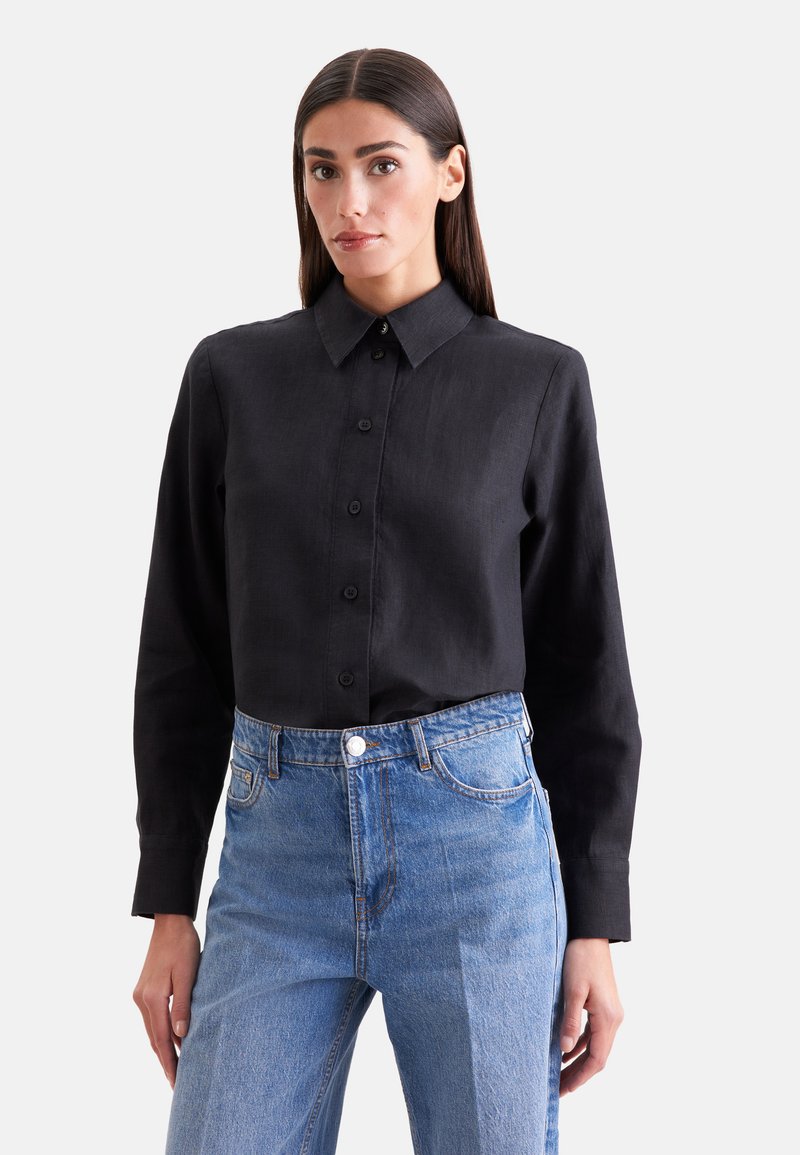 Femme portant une chemise noire boutonnée rentrée dans un jean bleu taille haute, debout les mains détendues le long du corps.