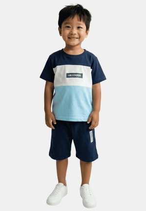 Garçon souriant debout, portant un t-shirt Lee Cooper bleu marine, blanc et bleu clair, un short bleu marine et des baskets blanches sur un fond uni.