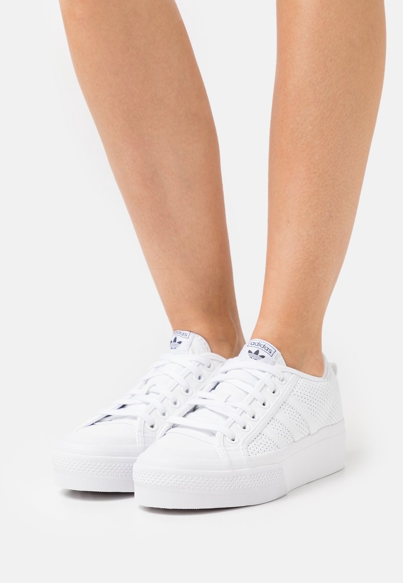 Vita sportskor med en perforerad överdel, platta snören och en tjock gummisula. Har tre ränder och en logotyp på tungan.