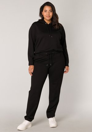Zwarte hoodie met een trekkoord en bijpassende joggingbroek. Zachte stof met een relaxte pasvorm en elastische enkels. Draagt met witte platform sneakers.