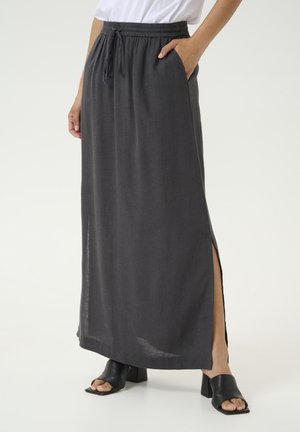 Vrouw in een lange donkergrijze rok met een split aan de zijkant, elastische taille met koord, wit topje en zwarte open-toe blokhakschoenen.