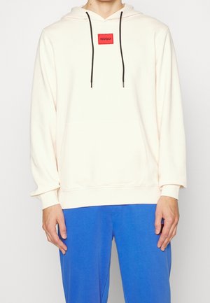Personne portant un sweat à capuche écru avec des cordons noirs et un écusson rouge avec le logo "HUGO", associé à un pantalon bleu vif.
