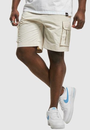 Beige Cargo-Shorts mit Seitentaschen, gestickten Akzenten und einer lockeren Passform, kombiniert mit weißen Sneakers mit hellblauen Details.