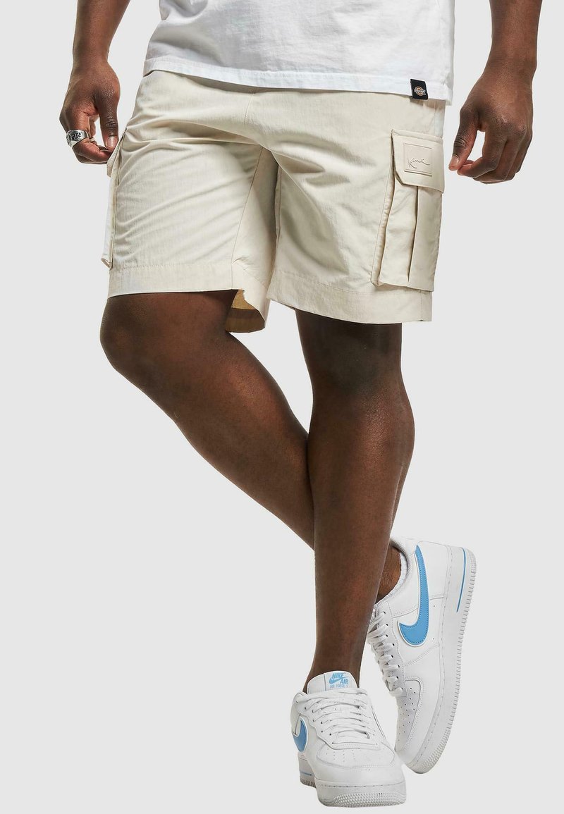 Beige cargo shorts med sidelommer, sydd detaljer og en avslappet passform, paret med hvite sneakers med lyseblå detaljer.