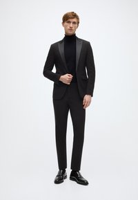 Zwart pak met satijnen revers, nauwsluitende zwarte coltrui, op maat gemaakte broek en gepolijste zwarte nette schoenen. Model staat tegen een witte achtergrond.