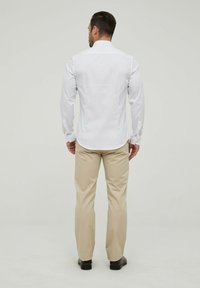Chemise blanche à manches longues avec un col classique, associée à un pantalon beige. La tenue présente une coupe ajustée et une texture lisse.