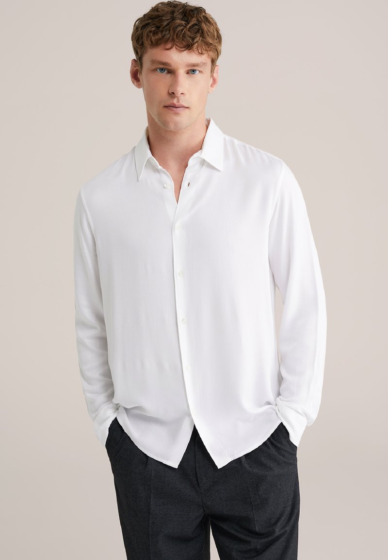 Biela polokošeľa s dlhým rukávom a mäkkou textúrou, s ostrým golierom a zakriveným lemom, nosená s tmavosivými nohavicami.