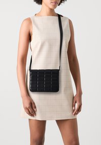 Bolso de hombro acolchado negro con forma rectangular, textura suave y una correa delgada ajustable, combinado con un vestido de cuadros claros.