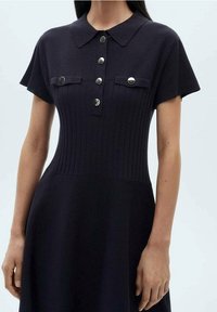 Robe en maille bleu marine foncé avec un corsage côtelé, des manches courtes, un col et des boutons métalliques à l'avant. Elle est dotée de deux poches poitrine.