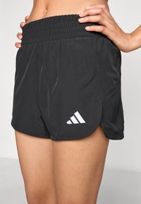 Svarta träningsshorts med en bred, rynkad midja, tillverkade av lätt material. Har vitt Adidas-logotyp och dubbellagersdesign.