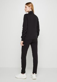 Conjunto de sudadera y pantalones de chándal negros en tejido suave. Presenta un cuello alto y puños con banda. Combinado con zapatillas blancas.