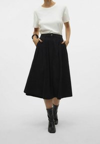 Jupe plissée noire en tissu texturé, longueur mi-mollet, assortie à un t-shirt blanc et des bottes noires montantes.