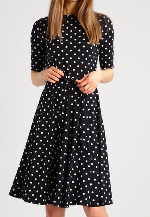 Femme portant une robe noire longueur genou à pois blancs avec des manches mi-longues, nouant une ceinture en tissu autour de la taille.