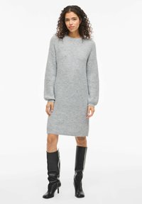 Graues gestricktes Kleid mit langen Ärmeln, rundem Halsausschnitt und knielangem Saum. Tragen mit knielangen schwarzen Stiefeln mit Absatz.