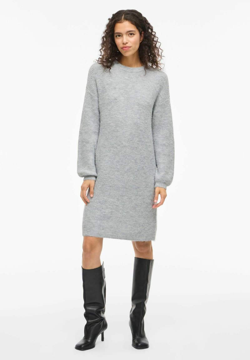 Graues gestricktes Kleid mit langen Ärmeln, rundem Halsausschnitt und knielangem Saum. Tragen mit knielangen schwarzen Stiefeln mit Absatz.