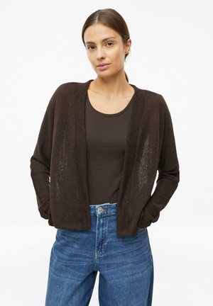 Frau trägt einen dunkelbraunen offenen Cardigan, ein dunkelbraunes Hemd und blaue hoch taillierte Jeans mit den Händen in den Taschen.