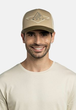 Mann lächelt, trägt eine beige und khakifarbene Netzkappe mit Berglogo und ein cremefarbenes Rundhalsshirt vor einfärbigem Hintergrund.