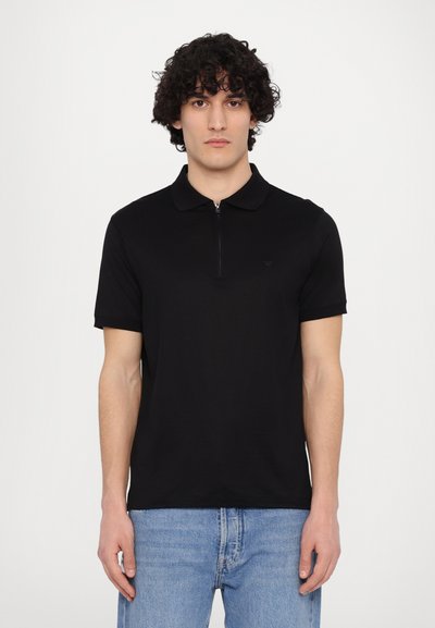 Black polo shirt with short sleeves, zipper collar, and a subtle logo detail on the chest. Pairing with light blue jeans.
