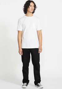 Biała bawełniana koszulka z kieszonką na piersi, zestawiona z czarnymi dżinsami i czarnymi butami do jazdy na deskorolce, stojąca na tle jednolitego tła.