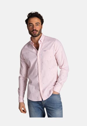 Mann trägt ein hellrosa gestreiftes Hemd mit kleinen Mustern, kombiniert mit blauen Jeans, steht vor einem einfarbigen Hintergrund.