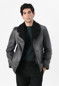 Giacca in shearling grigia con colletto nero texturizzato. Presenta una chiusura frontale con bottoni e una finitura liscia. Indossata sopra un maglione verde scuro.
