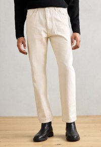 Pantalones de algodón beige con corte recto, bolsillos frontales y cierre de botón, combinados con botas de cuero negras hasta el tobillo sobre un suelo de madera.
