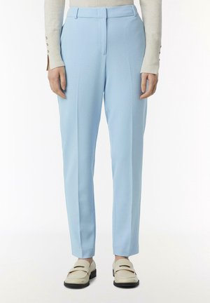 Personne portant un pantalon bleu clair sur mesure, un haut crème à manches longues avec des boutons sur les manches, et des chaussures crème à enfiler avec semelles noires.
