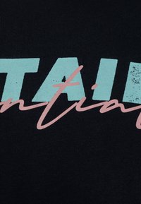 T-shirt noir avec de grandes lettres vert menthe épelant « STAIN » et un texte en cursif rose en dessous, avec une finition d'impression texturée.