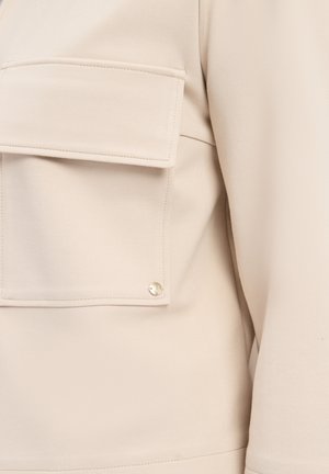 Vêtement beige avec une grande poche à rabat et des détails en métal doré. Tissu lisse avec une coupe structurée. Vue de côté mettant en avant le placement de la poche.
